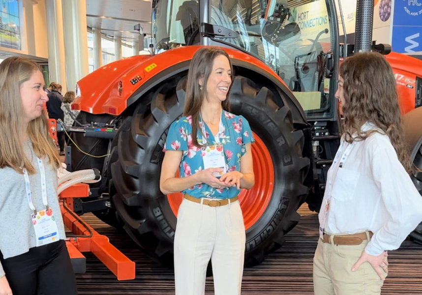 Heidi Wright standing in front of a red tractor wearing a floral blouse and white pants, smiling and talking with two other women.